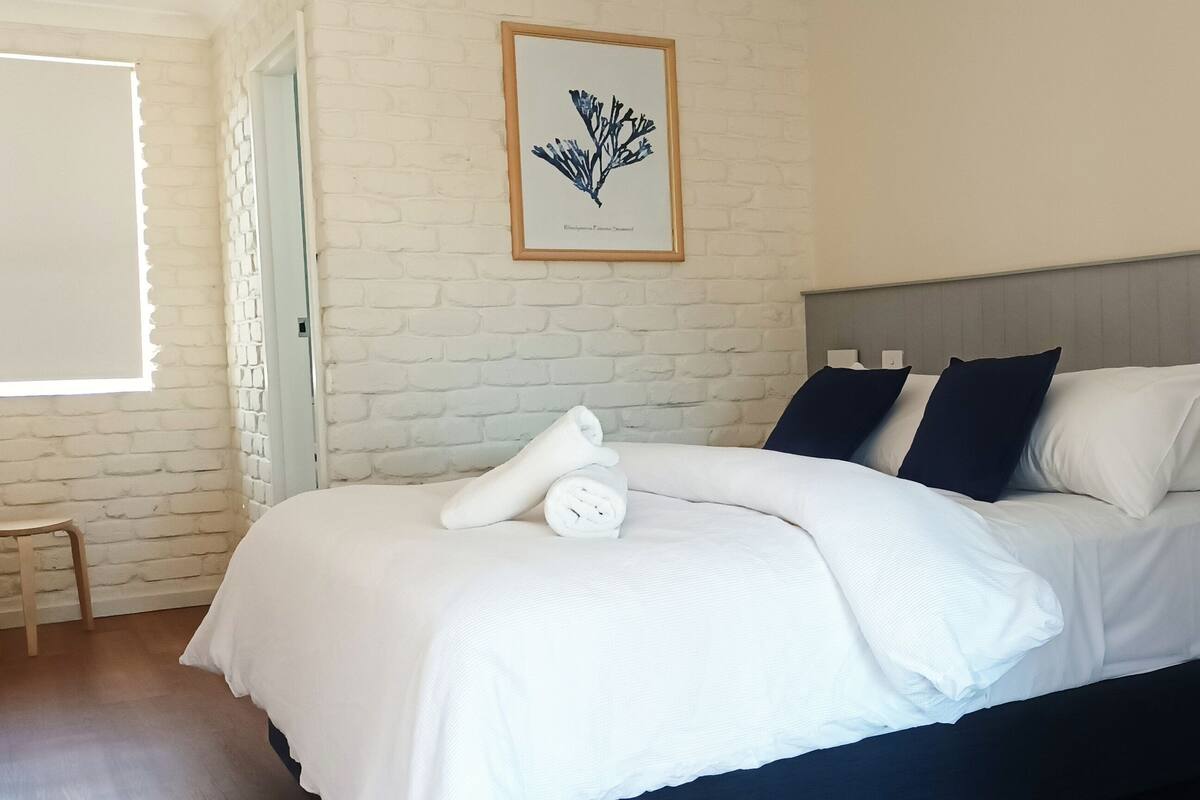 A neatly made bed with white linens and two pillows, a bedside table with a drawer, a framed botanical print on the wall, and a window with blinds.