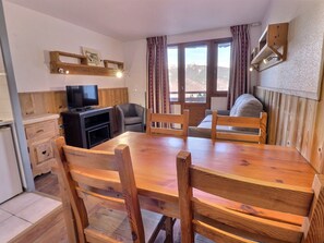 A wooden dining table with chairs, a television on a wooden cabinet, and a window with curtains.