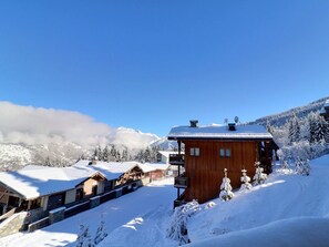 Un chalet en bois doté d’un balcon, entouré d’arbres couverts de neige et avec des montagnes en arrière-plan.