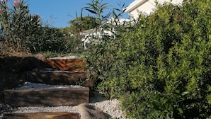 Um caminho de jardim com degraus de madeira e cascalho, rodeado por vegetação densa e um edifício branco ao fundo.