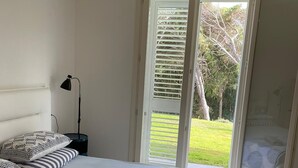 Quarto com cama, mesa de cabeceira, abajur e porta de vidro corrediça que dá para uma varanda.