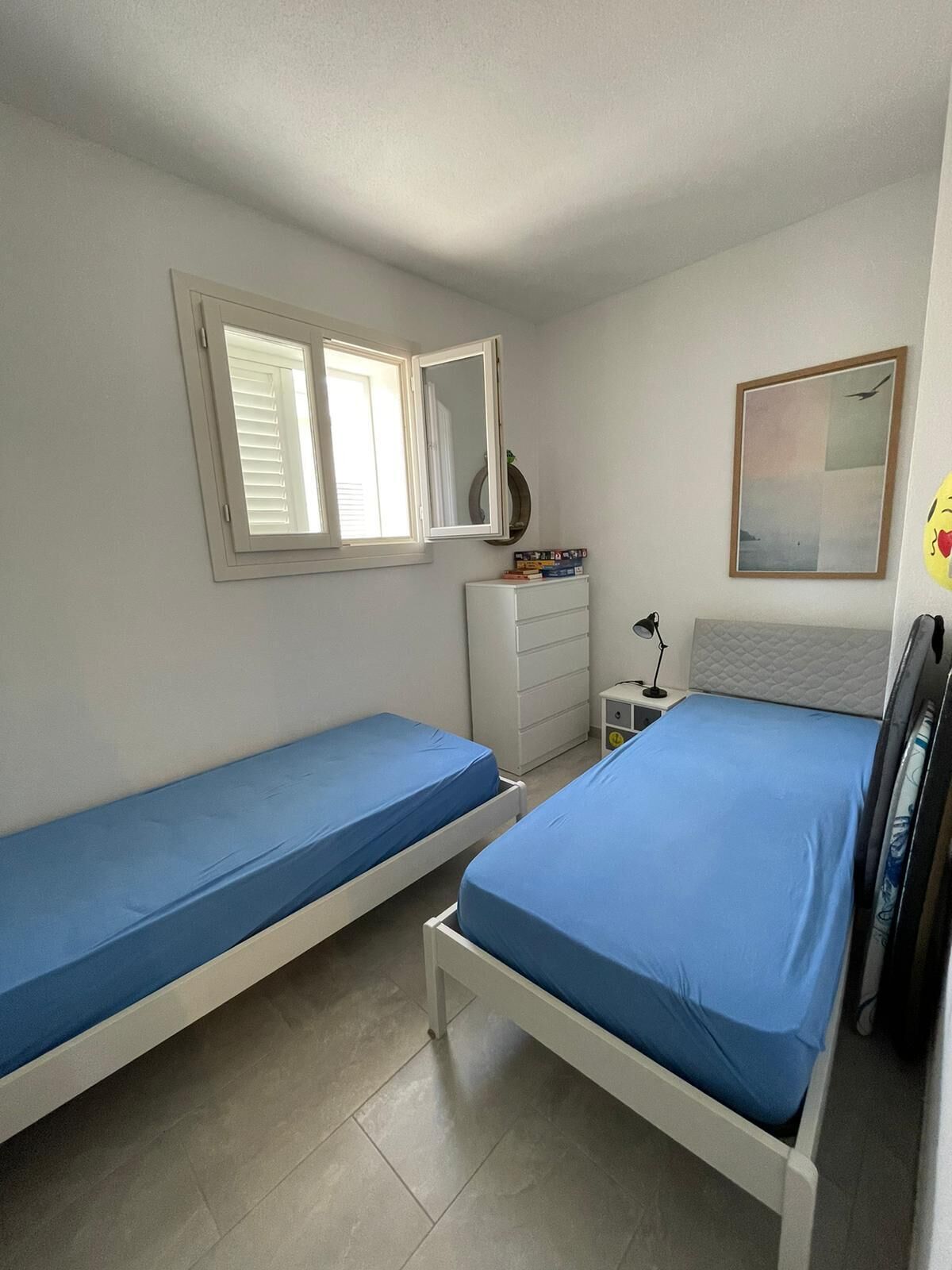 Ein Schlafzimmer mit zwei Einzelbetten, einem Schminktisch, einem Nachttisch mit Lampe, einem Fenster mit Jalousien und einem gerahmten Bild an der Wand.