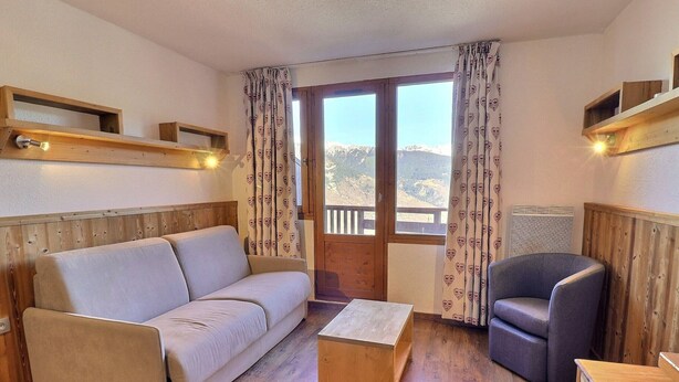 A cozy room with a sofa, a chair, a wooden coffee table, and a window with curtains. - Comfortable 2-Room with Balcony, Pets Allowed, Ski-in/Ski-out (La Tania)