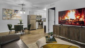 A modern living room with a sofa, a dining table with chairs, a kitchen area, and a large TV displaying a fireplace.