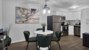 A modern kitchen with white cabinets, stainless steel appliances, a round dining table with green chairs, and a contemporary abstract painting on the wall.