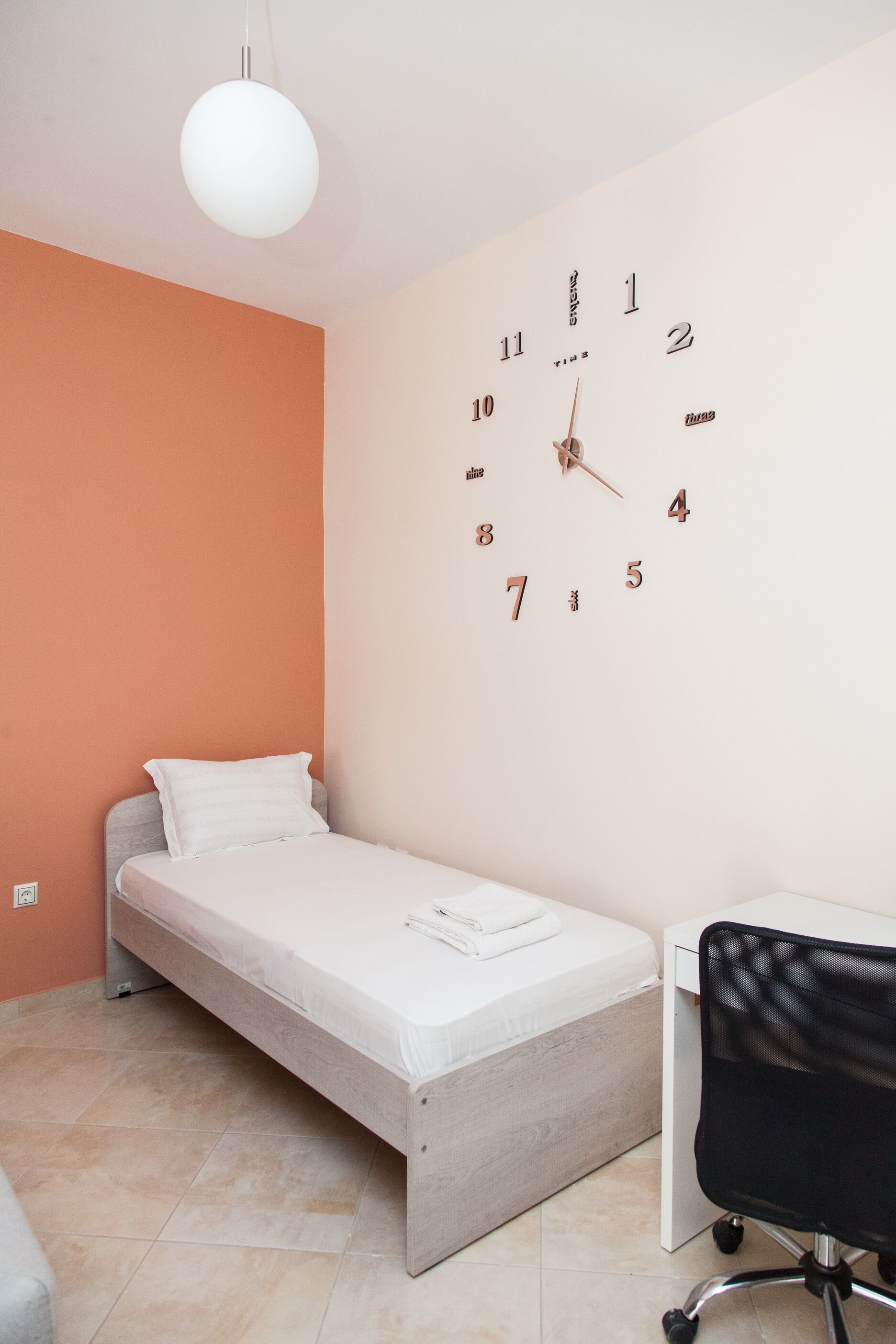 A small, modern bedroom with a single bed, a desk, and a wall clock.