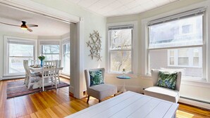 A bright, spacious room with wooden flooring, a dining area with white furniture, and large windows with blinds. - Dancing Queen by Bayley Vacation Rentals (Old Orchard Beach)