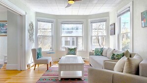 A spacious living room with a white ceiling fan, large windows, a white coffee table, and a beige sofa with patterned cushions. - Dancing Queen by Bayley Vacation Rentals (Old Orchard Beach)