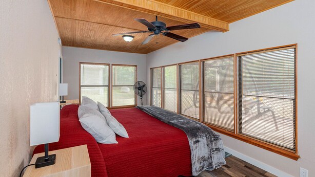 Une chambre avec un plafond en bois, un ventilateur de plafond, un lit recouvert d’une courtepointe rouge, une table de chevet avec une lampe et de grandes fenêtres équipées de stores.