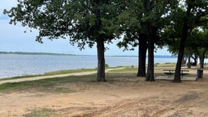 Un parc avec un sentier de terre, des arbres, un banc et une table de pique-nique situés près d’une étendue d’eau.