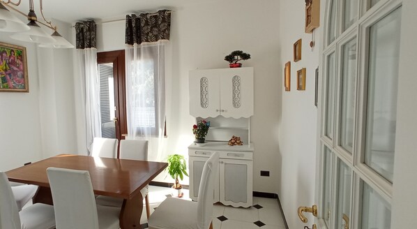 A dining area with a wooden table and white chairs, a white cabinet with decorative handles, and a window with sheer curtains.
