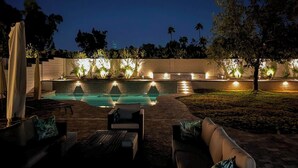 Zona de piscina exterior iluminada de noche, con sillones de descanso y un muro de piedra con luces.