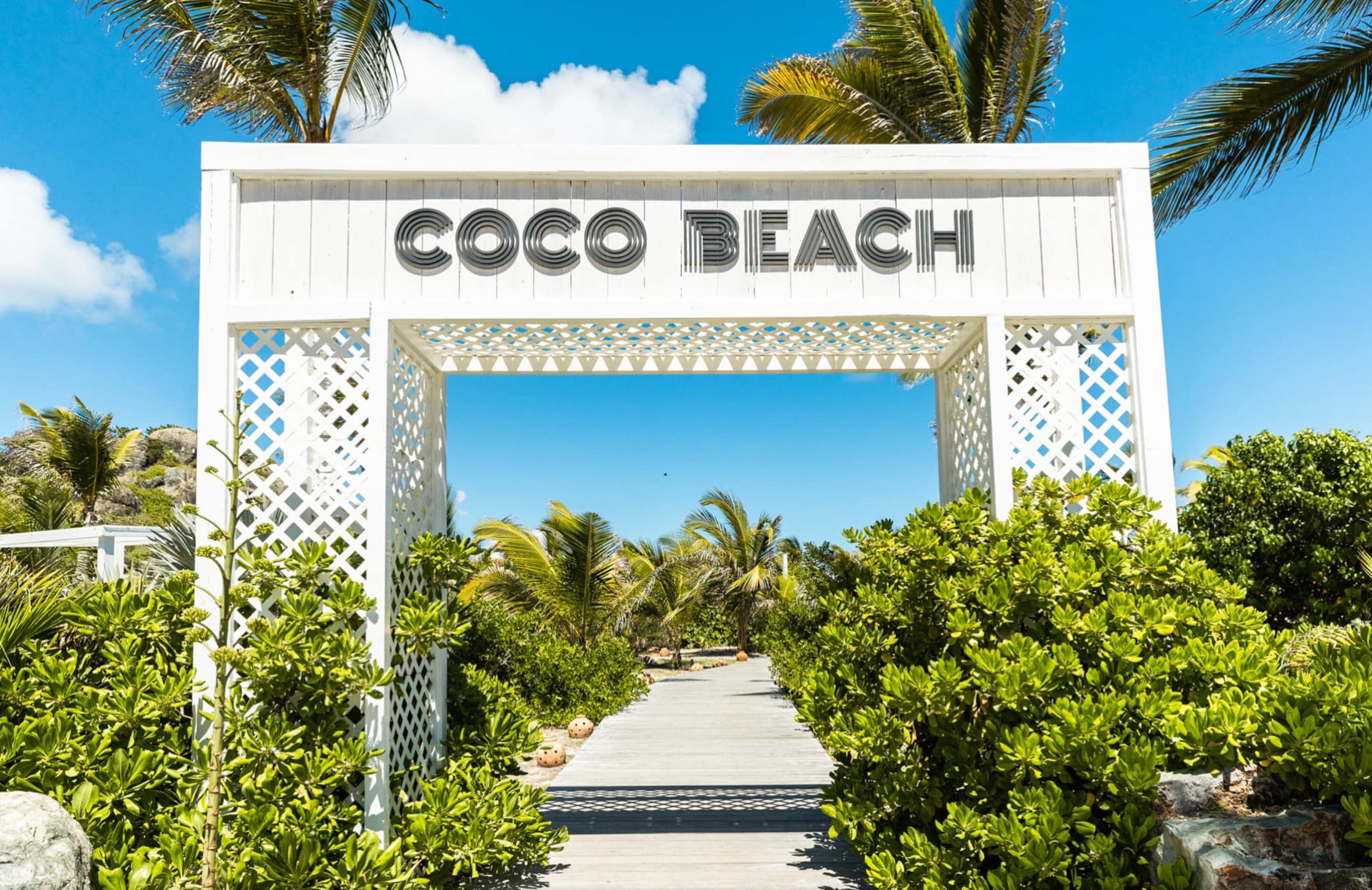 Valkoinen puinen holvikaari, jossa lukee teksti ”Coco Beach”, johtaa polulle, jonka varrella on vehreää kasvillisuutta ja palmuja.