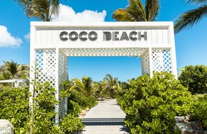 Un arco de madera blanca con el letrero "Coco Beach", que da acceso a un sendero rodeado de exuberante follaje y palmeras.