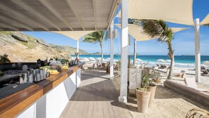 A beachside bar with a wooden counter, outdoor seating, and a view of the ocean.