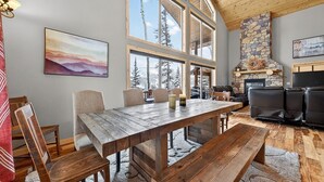 Amplia sala de estar con una gran mesa de madera, asientos cómodos, chimenea de piedra y ventanas grandes que ofrecen vistas a los árboles.