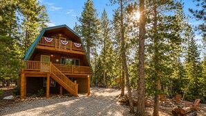 A cabin with a balcony, a staircase, and a gravel driveway surrounded by trees.