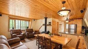 A cozy interior with a wooden ceiling, a dining area with a wooden table and chairs, a sofa with a moose pillow, and a kitchen with wooden cabinets and a stainless steel refrigerator.