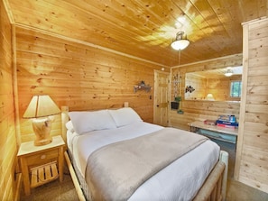 Una habitación de cabaña de madera con una cama, una mesita de noche con lámpara, un espejo y un estante con cajas.
