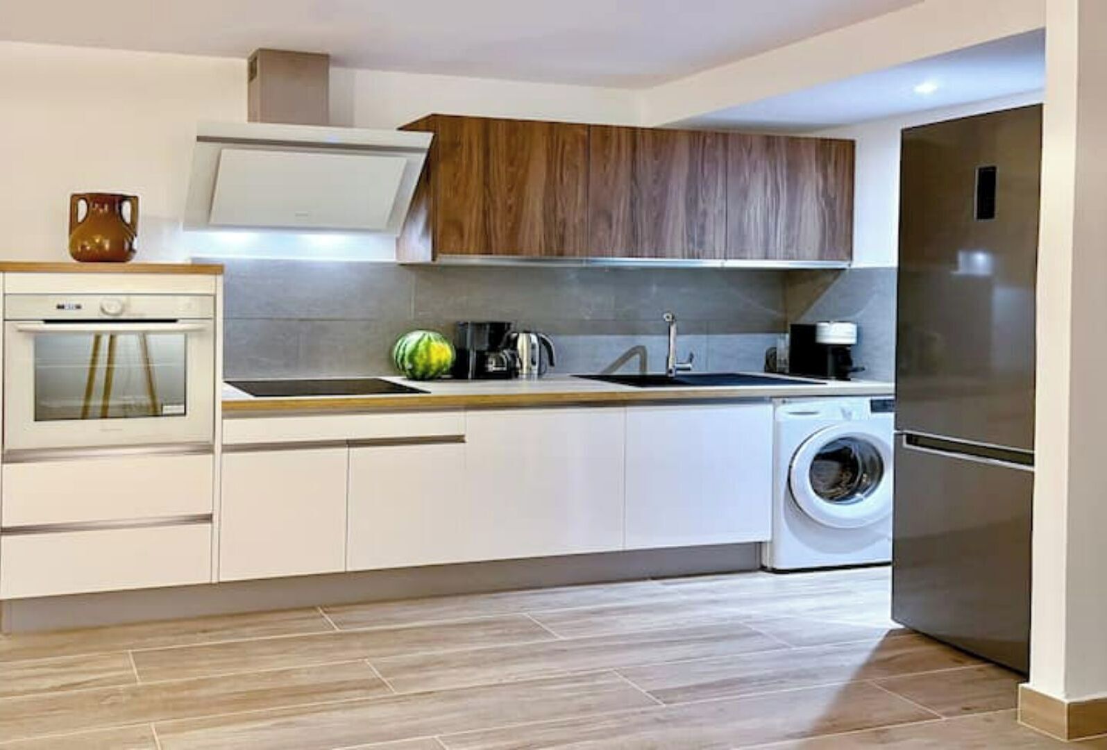 A modern kitchen with a built-in oven, a sink, a washing machine, and wooden cabinets.