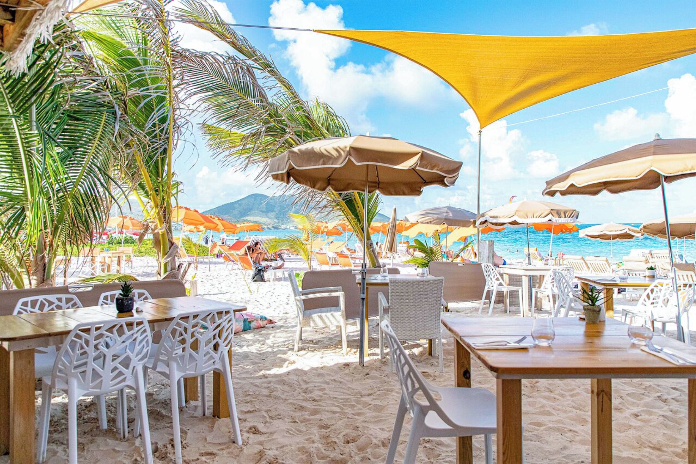A beachside restaurant with outdoor seating, including tables and chairs, under large umbrellas.