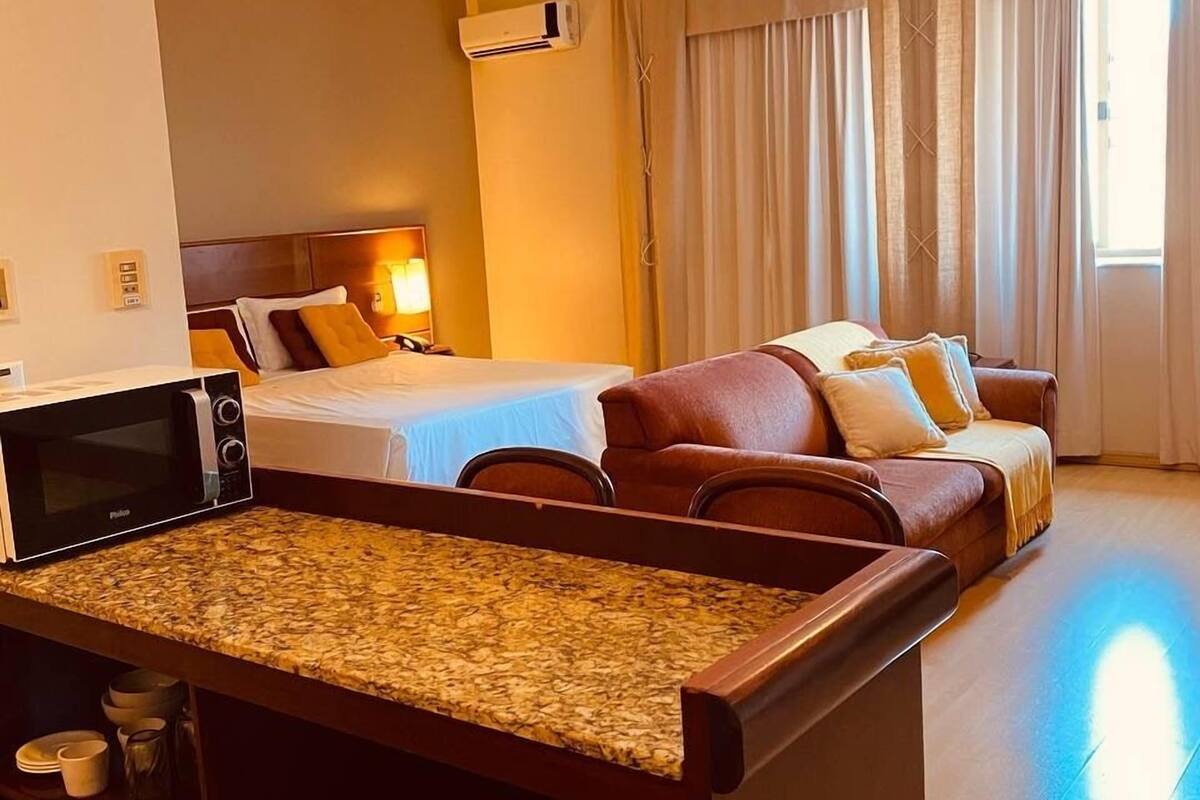 A hotel room with a kitchenette, a bed, a sofa, and a large window with curtains.