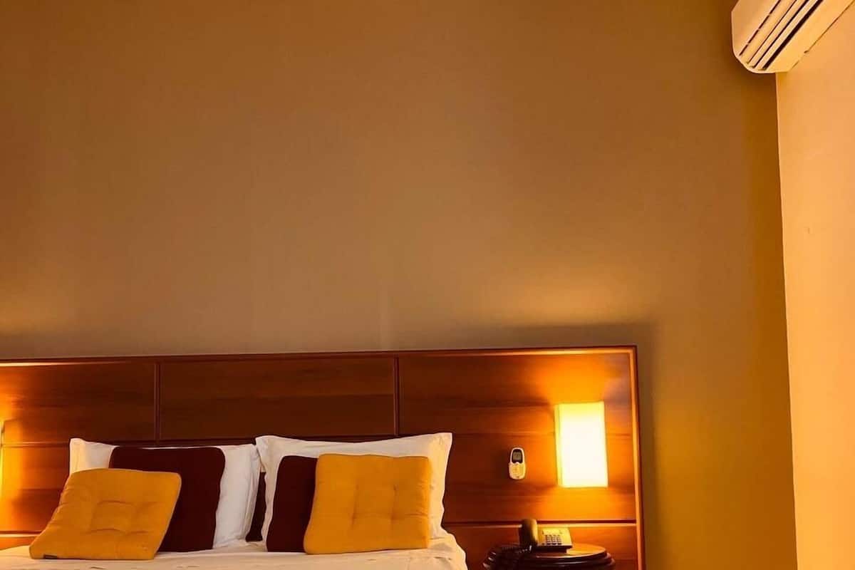 A hotel room with a bed, bedside table, lamp, and an air conditioning unit.