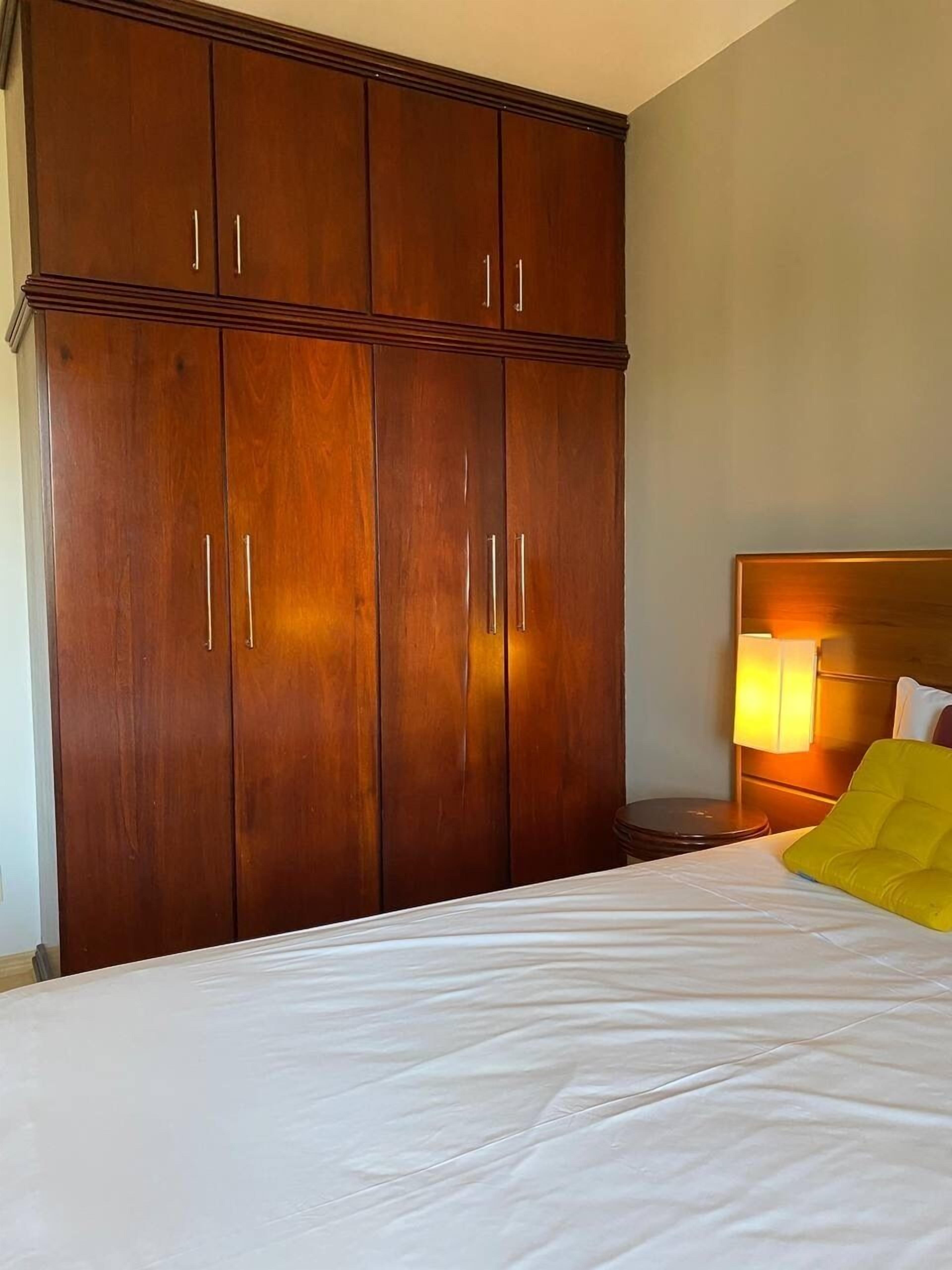 Habitación de hotel con un armario de madera grande, una cama con ropa blanca y una mesita de noche con lámpara.