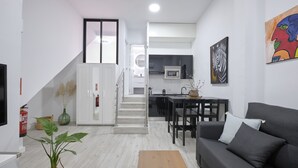 Un soggiorno moderno con un divano grigio, una zona pranzo con tavolo e sedie, una scala che conduce a un livello superiore e una cucina sullo sfondo.