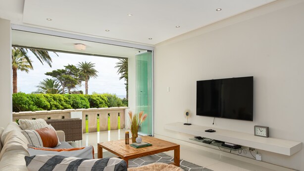 A modern living room with a sofa, coffee table, and TV. It features a balcony with a view of greenery and palm trees. - Sonnekus 5 (Cape Town)