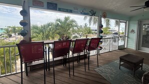 Área externa coberta com mesa e cadeiras, ventilador de teto e vista para palmeiras e edifícios.