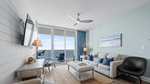 A spacious living room with a sofa, ottoman, and a dining area with a table and chairs. There is a ceiling fan, a large window with a view, and a painting on the wall. - Gulf Front Views | Indoor & Outdoor Pools, Hot Tub, Sauna, & Fitness Center (Gulf Shores)