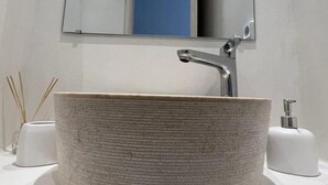 A bathroom with a round, textured sink, a mirror above it, and a blue door in the background. - Charming Apartment in Ano Mera (Mykonos)