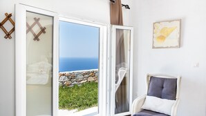 Una habitación con un gran ventanal corredera que da a vista al mar, una silla de mimbre con cojines y una obra de arte colgada en la pared.