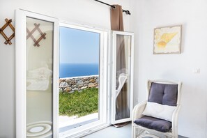 A room with a large sliding glass door leading to a view of the sea, a wicker chair with cushions, and a wall-mounted artwork.