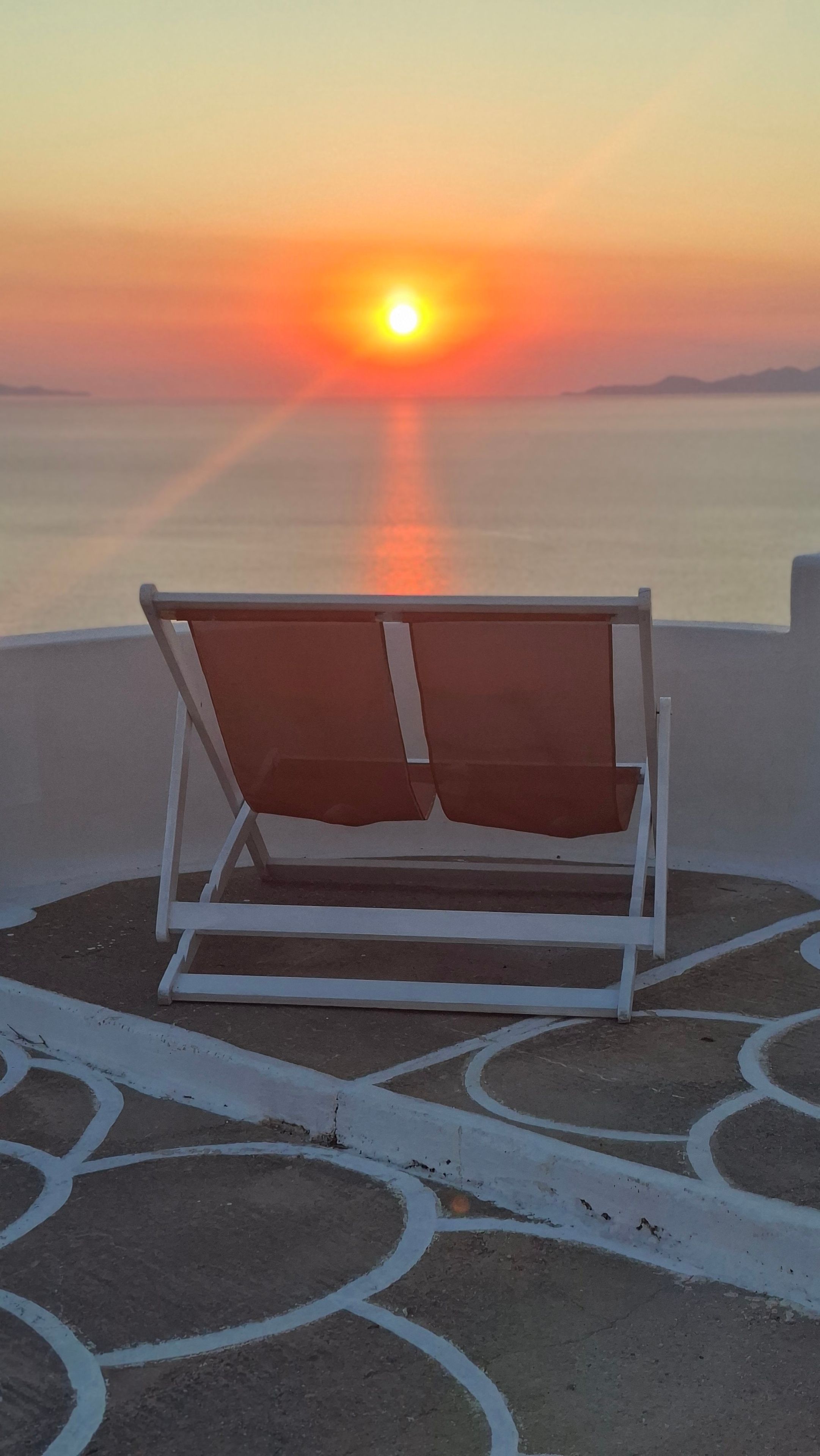 Una silla de descanso individual con cojín sobre un suelo exterior con diseño, con vistas a una masa de agua tranquila y un atardecer de fondo.