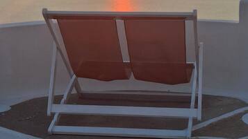 Una silla de descanso individual con cojín sobre un suelo exterior con diseño, con vistas a una masa de agua tranquila y un atardecer de fondo.