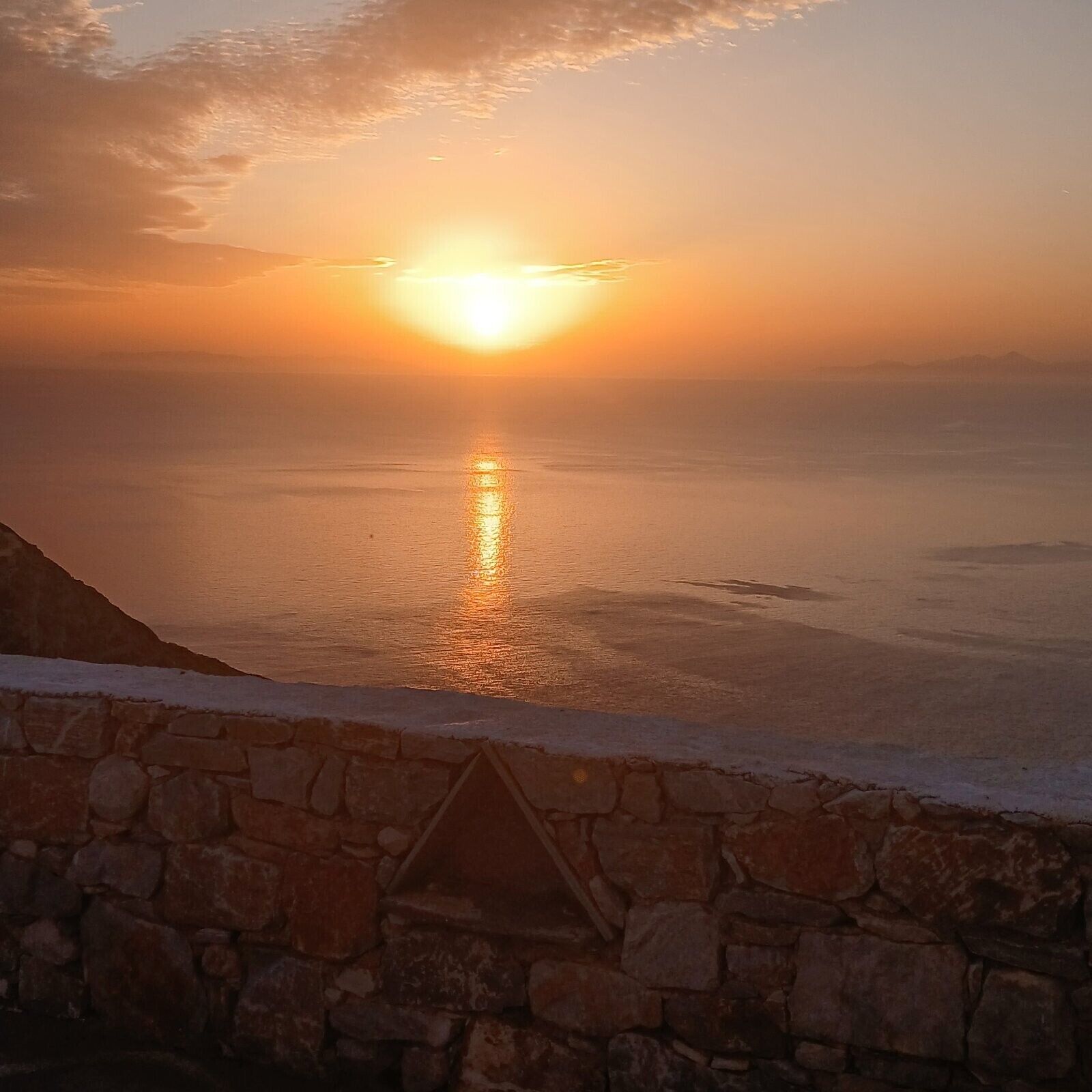 A sunset over a calm sea with a stone wall in the foreground.