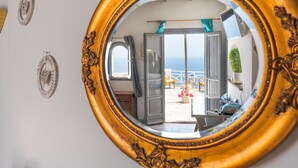 Un miroir rond, doté d’un cadre richement décoré, reflète une porte ouverte donnant sur un balcon d’où l’on peut admirer la mer.