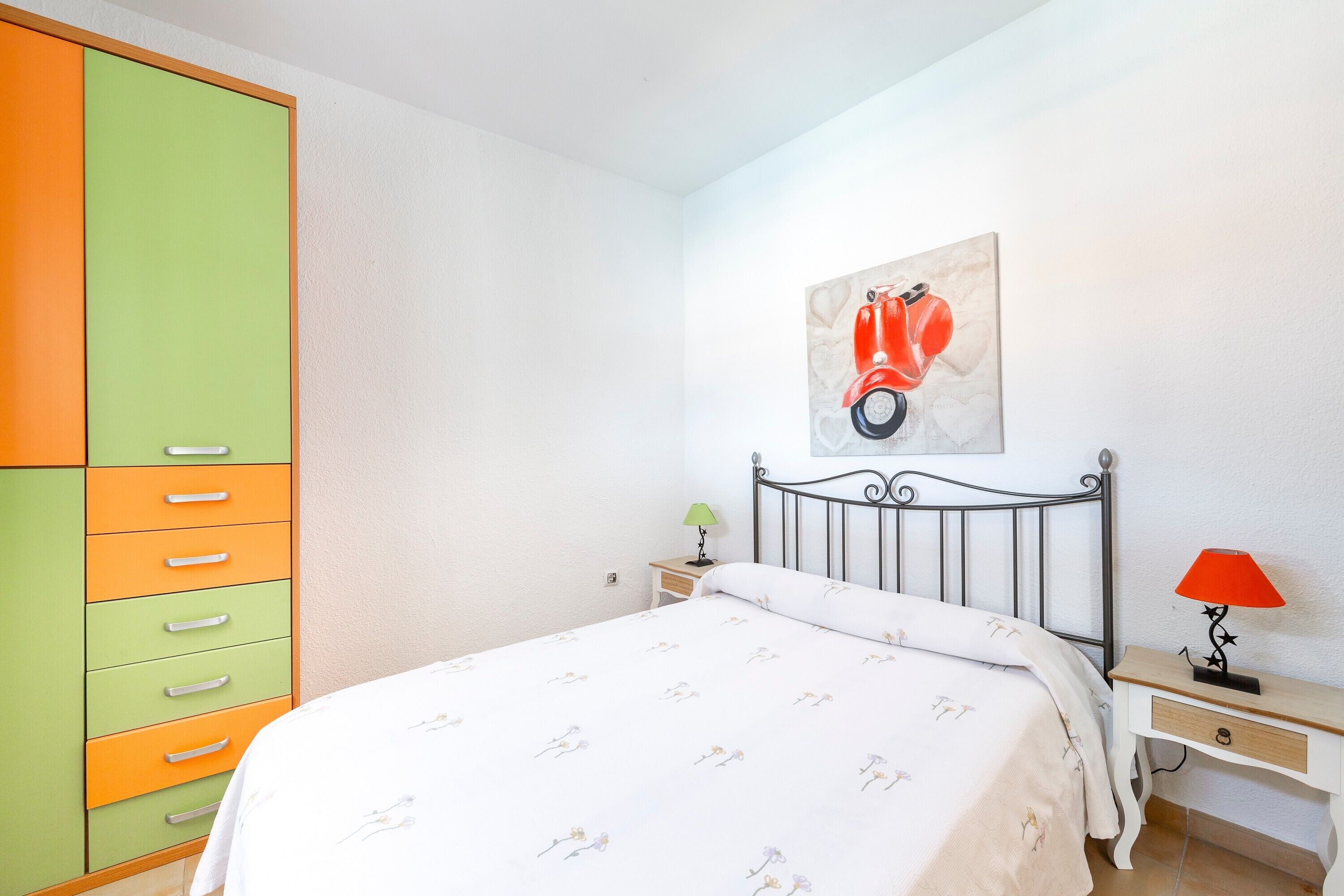 A bedroom with a bed, a nightstand, a lamp, a wardrobe, and a wall-mounted artwork.