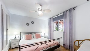 Ein Schlafzimmer mit einem Bett, einem Nachttisch, einem Deckenventilator, einem Fenster mit Vorhängen und einem Sessel.