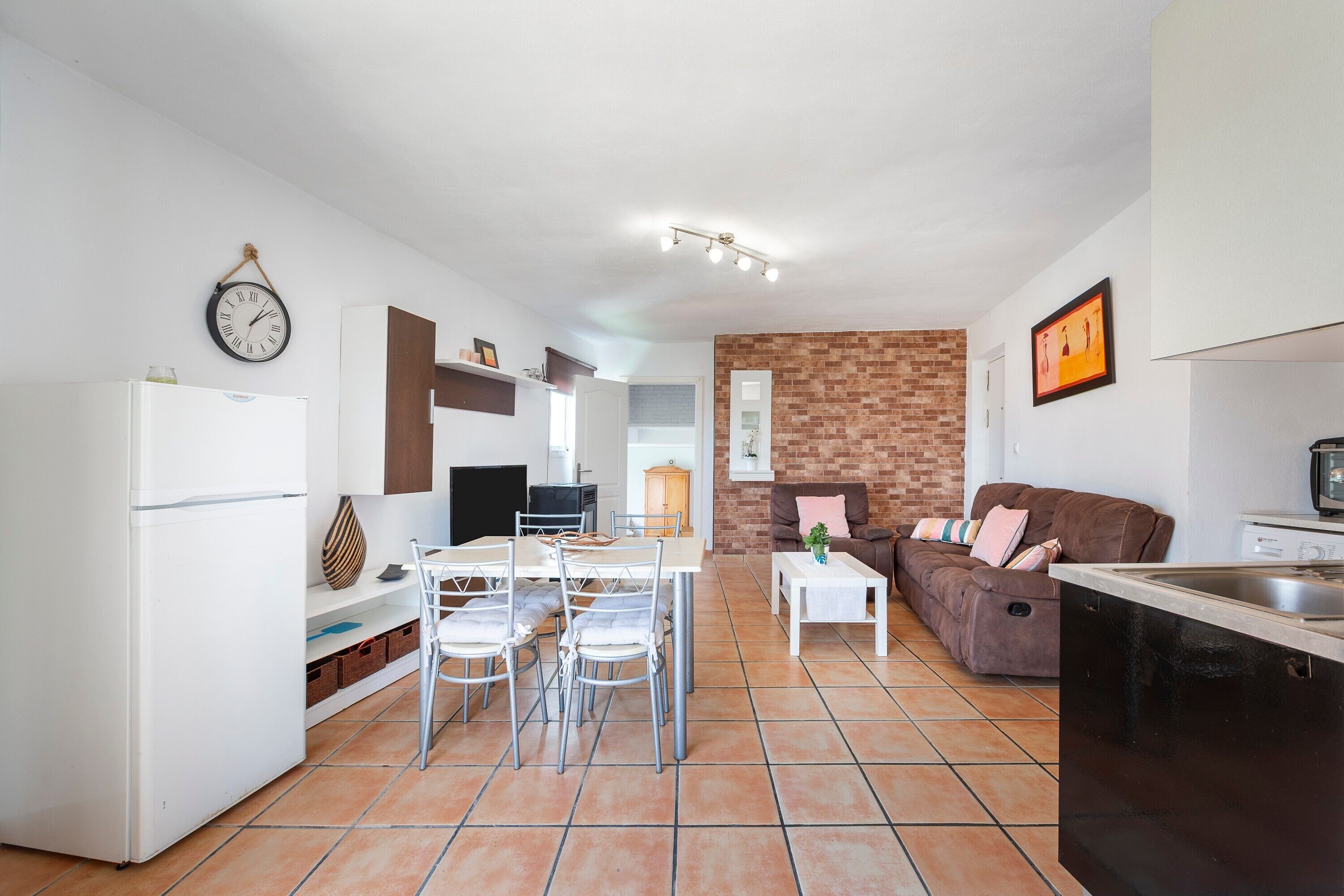 A modern kitchen with a refrigerator, dining table, and chairs, a living area with a sofa and coffee table, and a brick accent wall.