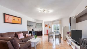 Un salon moderne avec un canapé en cuir marron, une table basse blanche, un coin repas avec une table et des chaises, une cuisine en arrière-plan et un téléviseur posé sur un meuble.