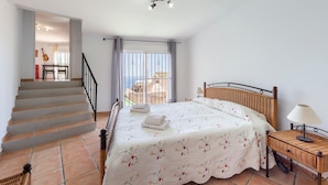 Une chambre à coucher avec un lit, une table de chevet, une lampe, une fenêtre avec des rideaux, un escalier et une vue sur l’extérieur.