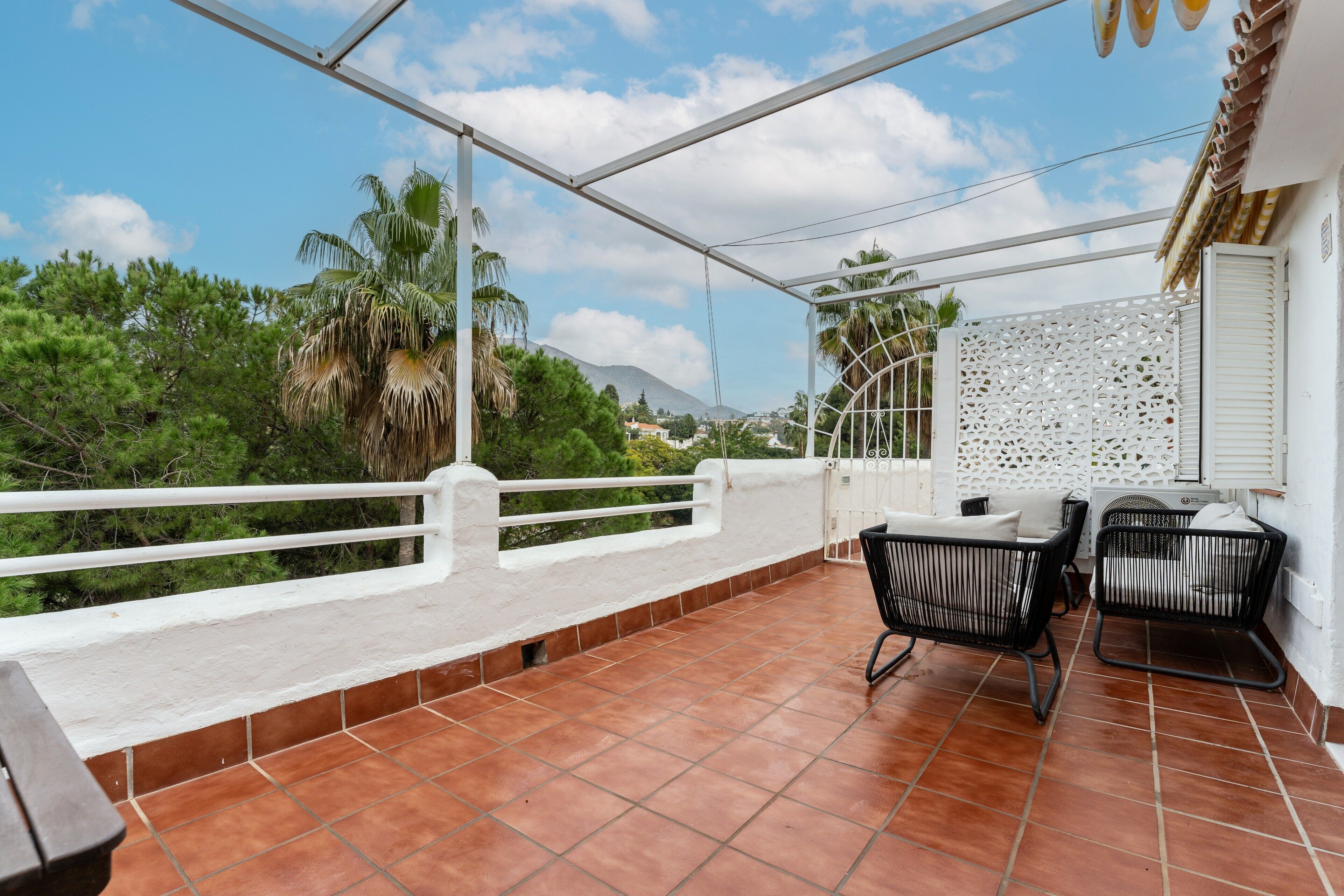 Una terraza techada con barandilla blanca, dos sillas negras y una pared blanca con un diseño decorativo.