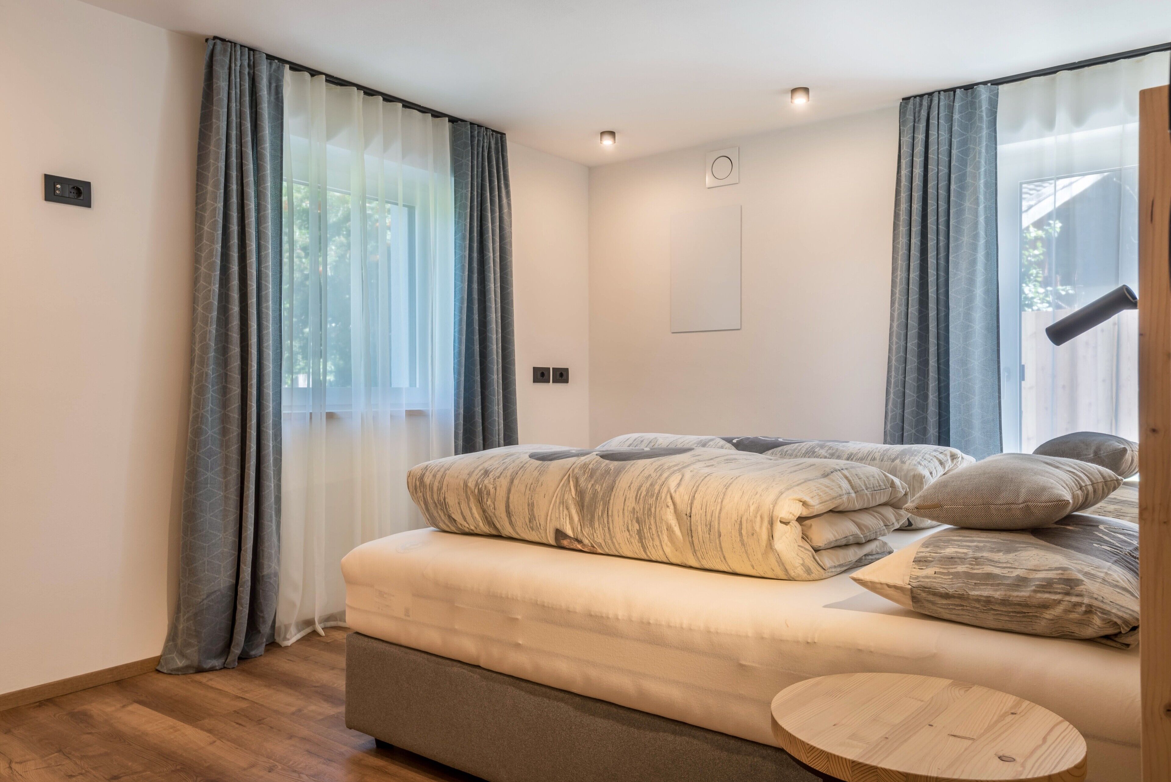 A modern bedroom with a bed, curtains, a round wooden side table, and a ceiling light.
