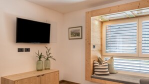 Habitació amb un televisor muntat a la paret, un armari de fusta, una banca amb coixins i una finestra amb persianes.