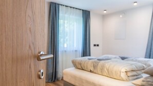 Un dormitori amb una porta de fusta, un llit amb matalàs i almoades grisos, una finestra amb cortines blaves i un interruptor a la paret.