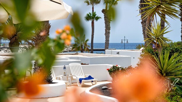 A poolside area with lounge chairs, a palm tree, and a view of the sea. - Vacation Home 'Puerto Chico B' with Sea View, Shared Pool & Wi-Fi (Playa Blanca)