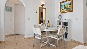A dining area with a round table, white chairs, a bookshelf, and a painting on the wall. - Vacation Home 'Puerto Chico B' with Sea View, Shared Pool & Wi-Fi (Playa Blanca)